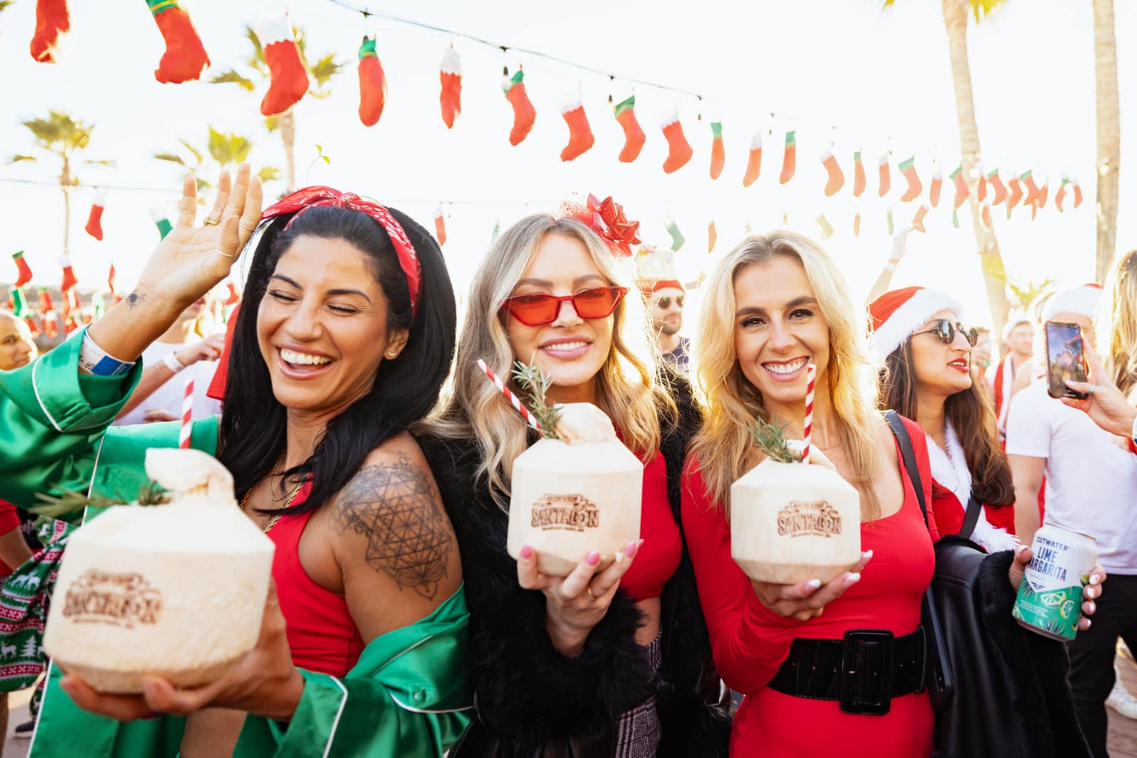 three women in Santa outfits holding branded coconuts at SantaCon at Belmont Park, under stockings strung from the lights