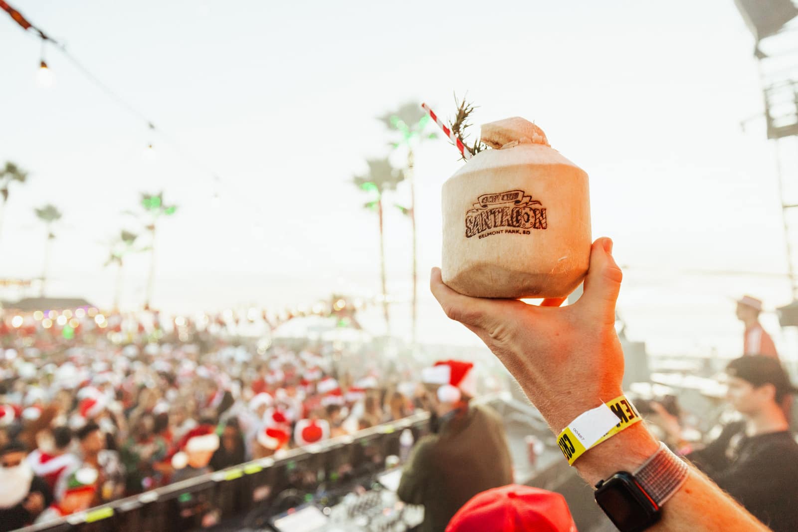 a hand holding a coconut hot-stamped with the Almost Nakey SantaCon Belmont Park logo above the crowd