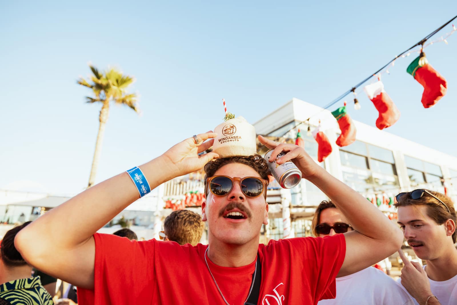 a man with a big mustache in a red tee balancing a stamped coconut on his head with a beer can in his other hand