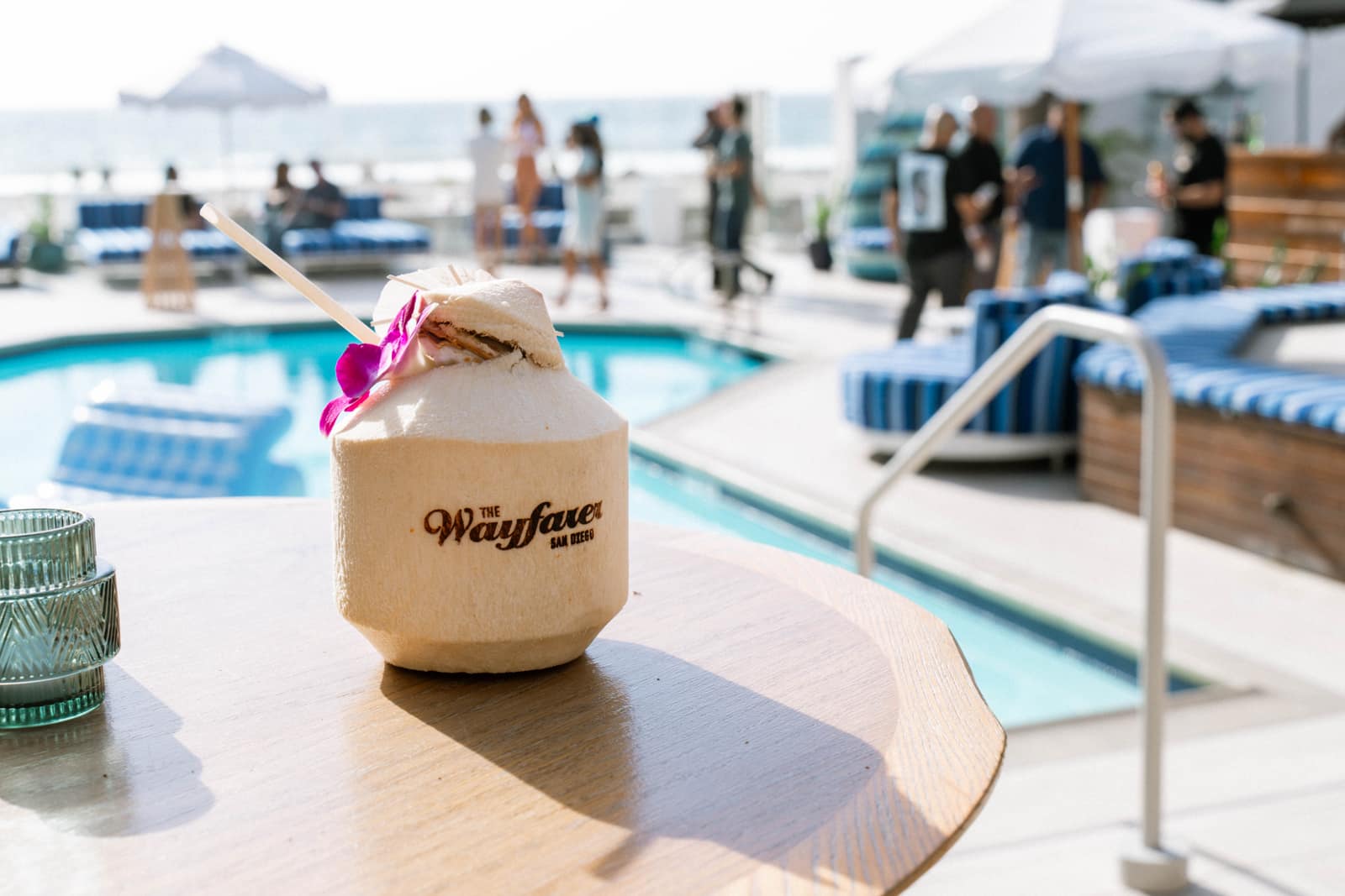 a custom coconut stamped with The Wayfarer San Diego logo, with a straw, a purple orchid and a paper umbrella, on a poolside table at a rooftop pool party
