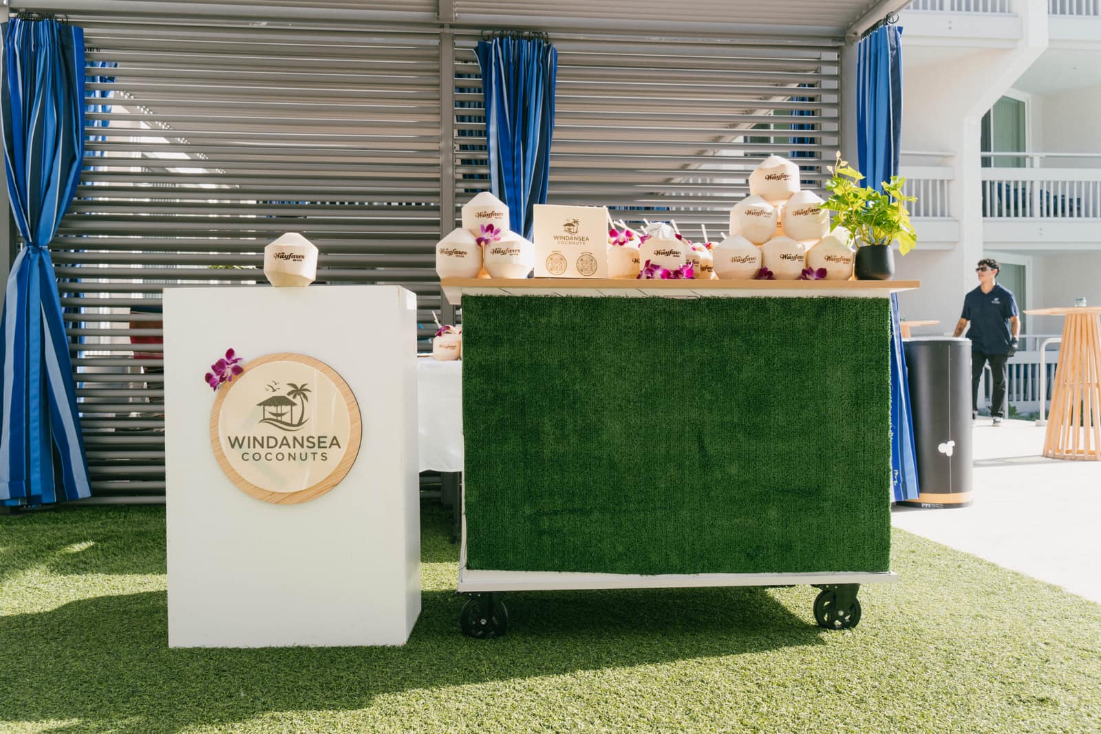 our coconut cart with a green turf front and coconuts stacked on top, next to a white podium with a round wooden sign with our logo